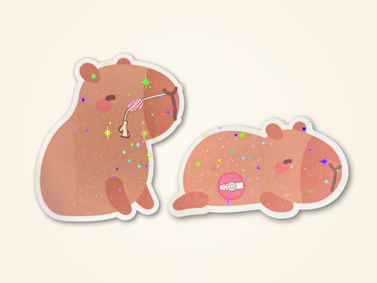 Two cute cartoon capybara stickers on a light cream background. One capybara is sitting and has a nasal feeding tube, and the other is lying down with a surgical feeding tube on its belly.