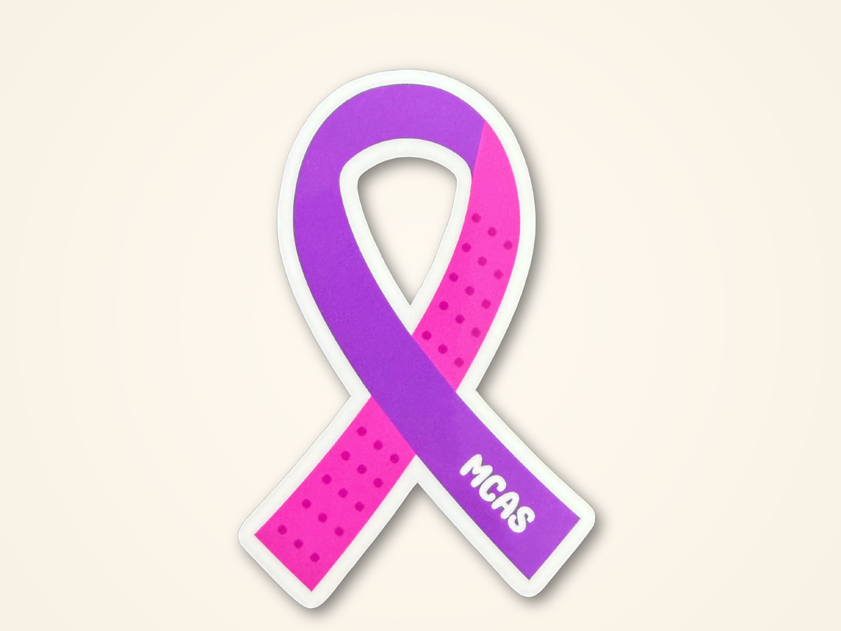 Awareness ribbon sticker in purple and pink with white dotted accents and the text 'MCAS' printed in white, shown on a light beige background.
