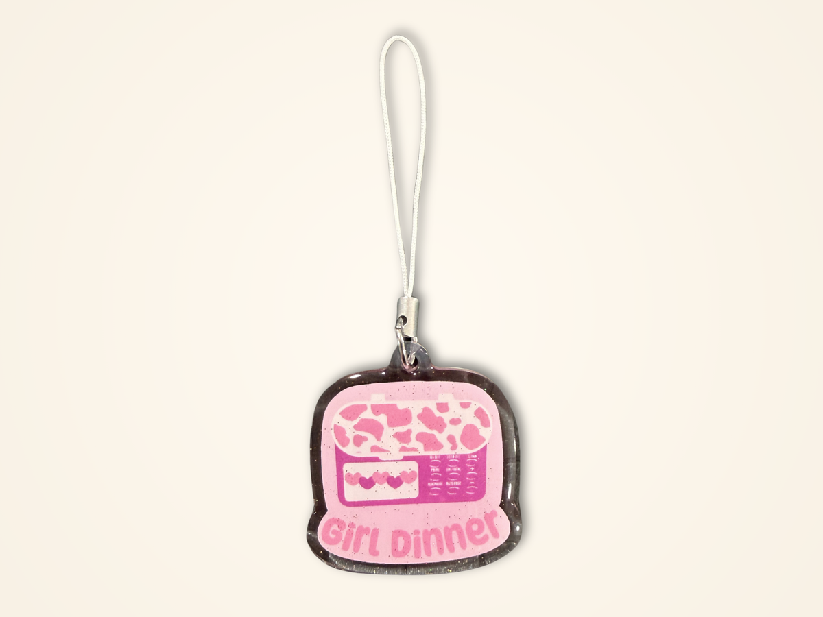 A small charm with a white loop strap, featuring a pink feeding tube pump illustration and the words 'girl dinner' on a light cream background.
