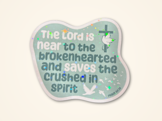 A sticker featuring the text 'The Lord is near to the brokenhearted and saves the crushed in spirit' from Psalm 34:14. The sticker has a light mint green background with dark teal lettering, white doves, a green cross, and subtle glitter accents, all set against a solid cream background.