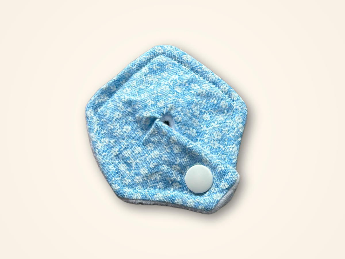 A light blue and white patterned reusable cloth pad with a white snap button, shown on a solid cream background.