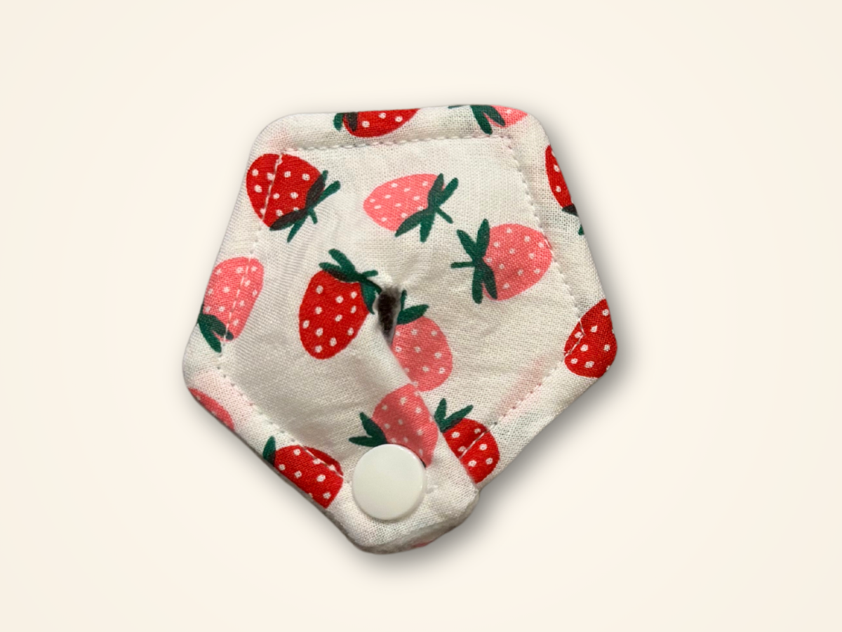 A hexagonal reusable cloth pad with a white background and a strawberry pattern, featuring a snap button closure at the bottom, shown on a light cream background.