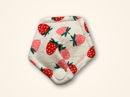 A hexagonal reusable cloth pad with a white background and a strawberry pattern, featuring a snap button closure at the bottom, shown on a light cream background.
